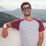 Young man in sunglasses leaning on a balcony with a scenic city view in the background.