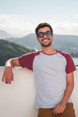 Young man in sunglasses leaning on a balcony with a scenic city view in the background.